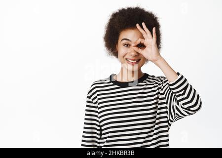 Porträt eines lächelnden afrikanischen Mädchens, das okay zeigt, mit null Geste auf dem Auge, auf weißem Hintergrund stehend Stockfoto