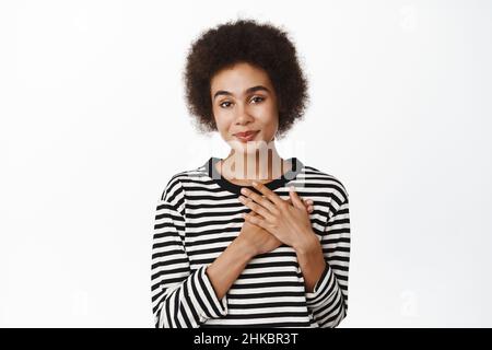 Beziehungs- und Valentinstag. Lächelndes afrikanisches Mädchen, das die Hände am Herzen hält und zufrieden, dankbar aussieht und auf weißem Hintergrund steht Stockfoto