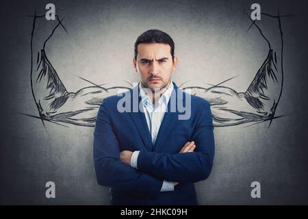 Porträt eines Geschäftsmannes, der gestreckt aussieht, isoliert auf einer grauen Wand mit einer Skizze muskulöser Arme, die hinter ihm Bizeps beugen. Wütende Person reagiert ärgerlich Stockfoto