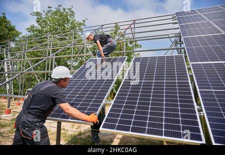 Männliche Arbeiter bauen Photovoltaik-Solarpanel-System im Freien. Ingenieure von Men platzieren Solarmodul auf Metallschienen, tragen Bauhelme und Arbeitshandschuhe. Erneuerbare und ökologische Energie. Stockfoto