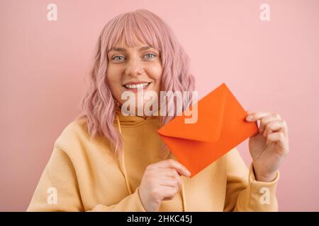 Porträt einer glücklichen 30s Frau, die einen roten romantischen Umschlag hält und die Kamera isoliert auf einem rosa Hintergrund anschaut. Valentinstag-Konzept. Stockfoto