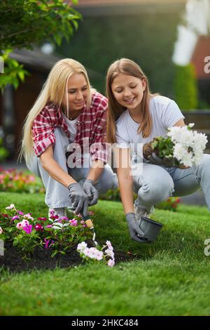 Angenehme junge Mutter, die mit ihrer charmanten Tochter Pflanzen auf dem Hinterhof eintopft. Teenager Mädchen hilft ihrer Mutter, um Blumen im Garten zu kümmern. Stockfoto