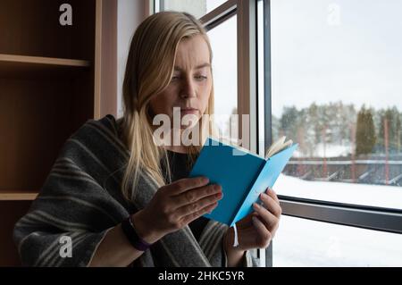 Frau, die ein gedrucktes Buch in sehr perieienbezügen liest und am Fenster mit Winterkulisse hinter sich steht Stockfoto