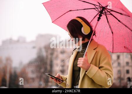 Junge Frau in Kopfhörern mit einem Regenschirm und Smartphone in der Stadt Stockfoto