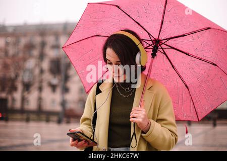 Junge Frau, die Smartphone mit Regenschirm in der Stadt hält Stockfoto