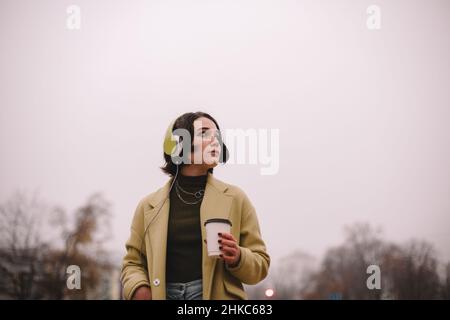 Nachdenkliche junge Frau, die in Kopfhörern Musik hört und durch die Stadt läuft Stockfoto