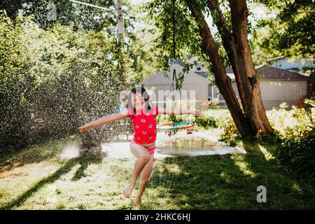 Im Sommer läuft das junge Mädchen durch den Sprinkler im Hinterhof Stockfoto