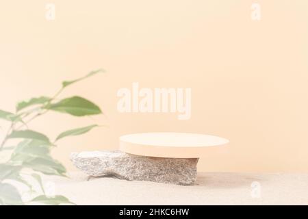 Rundes beigefarbenes Podium auf grünem Betonstein auf weißem Strandsand mit grünen Pflanzenblättern im Vordergrund. Kreative Komposition Hintergrund für Stockfoto