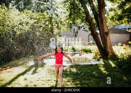 Im Sommer läuft das junge Mädchen durch den Sprinkler im Hinterhof Stockfoto