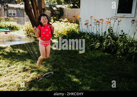 Im Sommer läuft das junge Mädchen durch den Sprinkler im Hinterhof Stockfoto