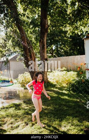 Im Sommer läuft das junge Mädchen durch den Sprinkler im Hinterhof Stockfoto