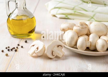 Rohe Champignons auf einem Teller und ein Glas Olivenöl auf einem weißen Holztisch. Kochen von frischen Champignons Agaricus bisporus. Konzept der vegetarischen Küche. Stockfoto