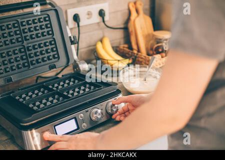 Die junge Frau hat Waffeleisen dabei und bereitet belgische Waffeln in der Küche zu. Kochvorgang. Stockfoto