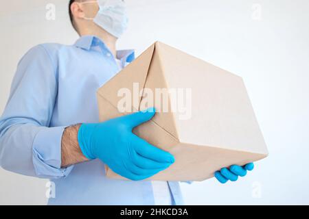 Der Lieferer hält einen Karton in einer medizinischen Maske und medizinischen Handschuhen. Express-Lieferung, Online-Shopping. Spendenbox für Coronavirus. Lieferung von Lebensmitteln Stockfoto