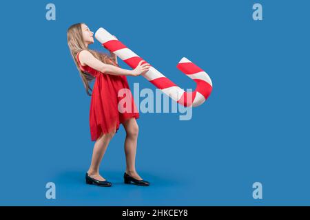 Ein wunderschönes kleines Mädchen, das auf blauem Hintergrund eine riesige Süßigkeit in den Händen hält. Weihnachtliche Süße. Foto-Session zum neuen Jahr. Großes Karamell. Teenager Stockfoto