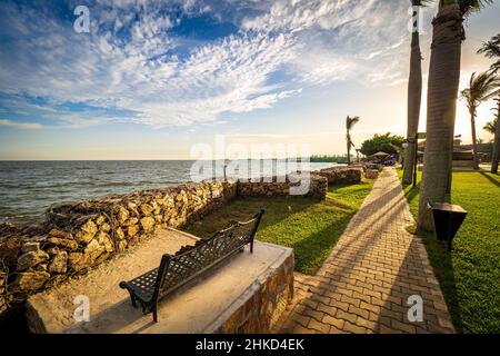 Blick auf den Gehweg und eine schmiedeeiserne Parkbank am Ufer des Viktoriasees in Entebbe, Uganda, bei den letzten Sonnenstrahlen am Abend Stockfoto