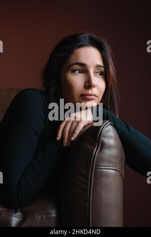 Junge attraktive nachdenkliche Frau mit langen dunklen Haaren in schwarzem Rollhalsausschnitt sitzen auf braunem Sofa und setzen den Kopf auf gekreuzte Arme. Stockfoto