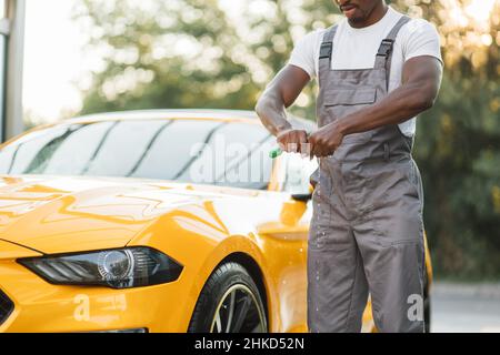 Nahaufnahme eines hübschen bärtigen jungen afrikanischen Arbeiters, der einen Lappen auf einem Hintergrund eines modernen gelben Luxusautos entschrauben kann. Ein Mann, der Auto mit einem Mikrofasertuch putzt. Der Mann hält die Mikrofaser in der Hand Stockfoto