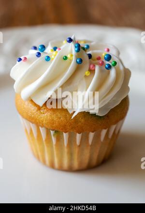 Eine Nahaufnahme eines Mini-Cupcake, der mit Perlmutt-Süßigkeitenkugeln verziert ist. Konfetti-Cupcake-Teig mit weißem Zuckerguss auf einem weißen Teller. Holztisch verschwommen Stockfoto