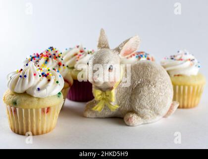 Kleine, frühlingshafte Cupcakes, die um eine Hasenfigur angeordnet sind. Konzentrieren Sie sich auf die vordersten Cupcakes mit zusätzlichen Unschärfen im Hintergrund. Weißer Hintergrund Stockfoto