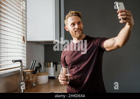 Attraktiver, fröhlicher junger Mann, der zu Hause ein Selfie macht, während er Wasser aus einem Glas trinkt Stockfoto