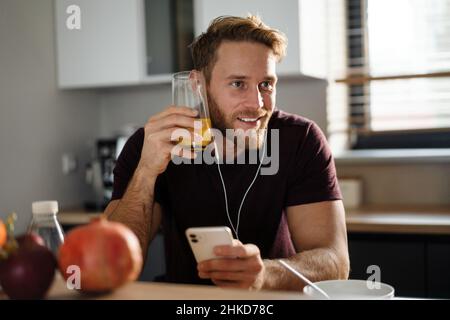 Schöner junger Mann, der mit Kopfhörern Musik hört, auf dem Mobiltelefon stöbert, während er in der Küche sitzt und Apfel isst Stockfoto