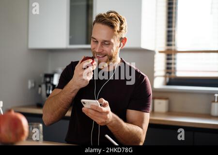 Schöner junger Mann, der mit Kopfhörern Musik hört, auf dem Mobiltelefon stöbert, während er in der Küche sitzt und Apfel isst Stockfoto