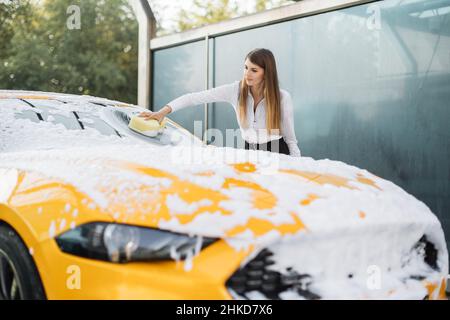 Horizontale Ansicht der Autoreinigung bei der Selbstwäsche im Freien. Eine Geschäftsfrau trägt ein weißes Hemd und eine schwarze Hose im Anzug, die ihre Windschutzscheibe mit gelbem Schwamm und Schaum wäscht. Stockfoto