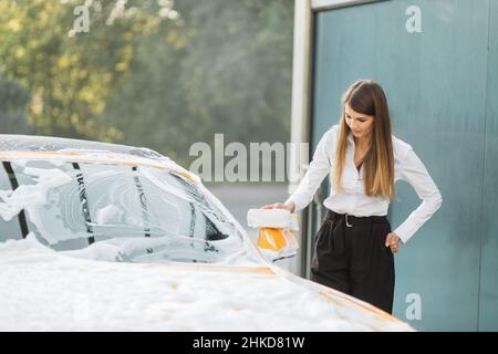 Horizontale Ansicht der Autoreinigung beim Selbstwaschservice. Kaukasische Geschäftsfrau trägt ein weißes Hemd und eine schwarze Hose, während sie den Rückspiegel mit Schwamm und Schaumstoff wäscht Stockfoto