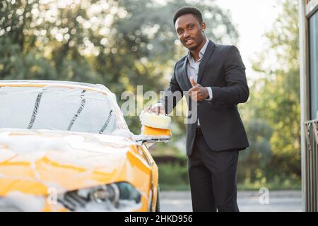 Horizontale Ansicht der Autoreinigung beim Selbstwaschservice. Hübscher, stilvoller afrikanischer Geschäftsmann, der mit einem glücklichen Lächeln auf die Kamera blickt, während er den Rückspiegel mit Schwamm und Schaumstoff wäscht, der den Daumen nach oben zeigt Stockfoto