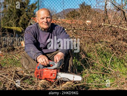 Glücklicher älterer Mann mit Kettensäge, der nach dem Schneiden des Baumes im Garten zur Ruhe sitzt Stockfoto