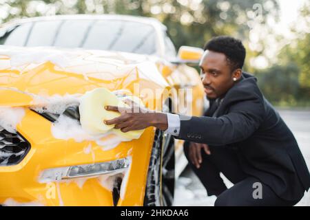 Junger gutaussehender bärtiger afrikanischer Geschäftsmann in formeller Kleidung, der seinen Luxus-Sportwagen im Freien an der Waschstation für Selbstwagen mit gelbem Lappen und Seifenschaum reinigte Stockfoto