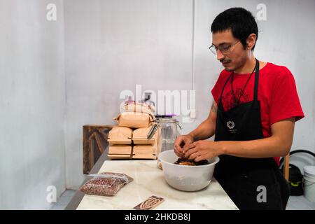 Ein mexikanischer Mann fügt Zimt in zerkleinerte Kakaobohnen vor dem Mahlprozess in der handwerklichen Schokoladenherstellung in Xochistlahuaca, Mexiko, hinzu. Stockfoto