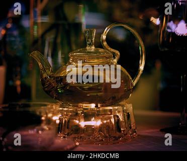 Transparente Teekannen mit grünem Tee auf einem beheizten Glasständer auf einem Restauranttisch mit reflektierenden Lichtstrahlen Stockfoto