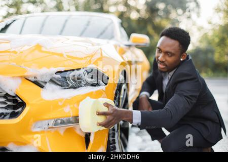 Seitenansicht des jungen, hübschen bärtigen, eleganten afrikanischen Geschäftsmannes, der sein gelbes Luxusauto im Freien an der Autowaschstation mit gelbem Lappen und Seifenschaum reinigte Stockfoto