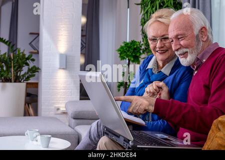 Ein reizendes Seniorenpaar sitzt auf einem Sofa und unterhält sich mit ihren Kindern und Enkelkindern per Videoanruf auf einem Laptop in ihrem Wohnzimmer. Stockfoto