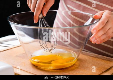 Nahaufnahme der Hände einer Frau, die Eier schlagen, um Teig auf dem Holztisch in der Küche zu machen. Hausgemachte Küche. Stockfoto