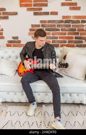 Junger Mann spielt E-Gitarre. Lernt Gitarre zu spielen. Interessantes Hobby. Weißes Sofa im Zimmer. Vertikal. Stockfoto