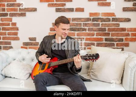 Junger Mann spielt E-Gitarre. Lernt Gitarre zu spielen. Interessantes Hobby. Weißes Sofa im Zimmer. Angenehme Erholung. Stockfoto