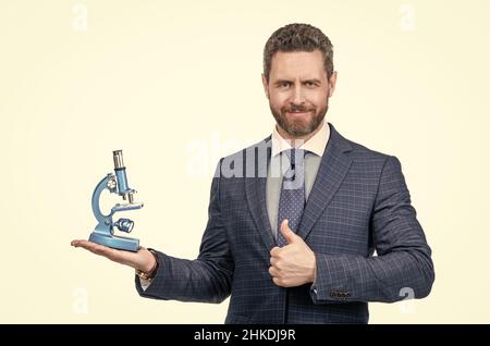 Mann im Anzug präsentiert moderne Mikroskop isoliert auf weißem zeigen Daumen nach oben, Wissenschaft und Wirtschaft. Stockfoto