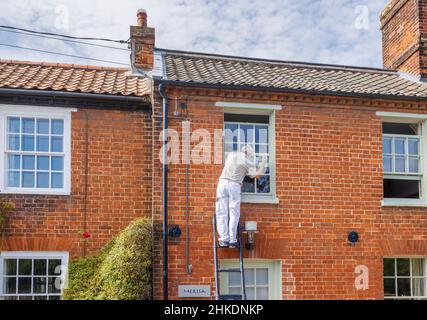 Ein älterer Mann auf einer Leiter malt den Fensterrahmen eines Hauses in Cley-Next-the-Sea, einem Dorf an der Nordküste von Norfolk, East Anglia, England Stockfoto