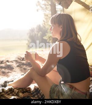 Kaffee und Kontemplation im Camp. Aufnahme einer jungen Frau, die während des Campens in einem Zelt sitzt und Kaffee trinkt. Stockfoto