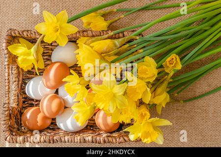 Ein Strauß Narzissen und ein Korb mit frischen weißen und braunen Eiern schließen sich auf rustikalem Hintergrund direkt von oben an Stockfoto