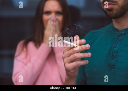 Passivrauchen. Mann raucht Zigarette in der Nähe einer Frau, die ihr Gesicht vor Zigarettenrauch bedeckt. Stockfoto