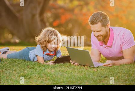 Porträt eines Mannes mit Sohn, der draußen ein digitales Tablet benutzt. Neue normale wieder in die Schule. Schüler des Elementargelehrten, der mit dem Lehrer auf Gras sitzt. Stockfoto