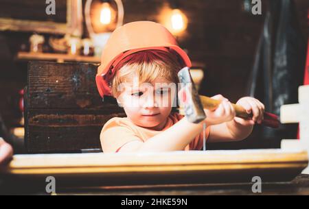 Kid hämmert Nägel mit einem Hammer in ein Holzbrett. Junge Kind beschäftigt im Helm lernen, Hammer zu verwenden. Kind hämmert Nagel in Holz zu Hause. Stockfoto