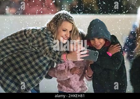 Prattville, Alabama, USA- 23. Dezember 2021: Eine Frau posiert mit einem jungen Mädchen und Jungen im künstlichen Schnee, während sie am Telefon ein Selfie macht Stockfoto