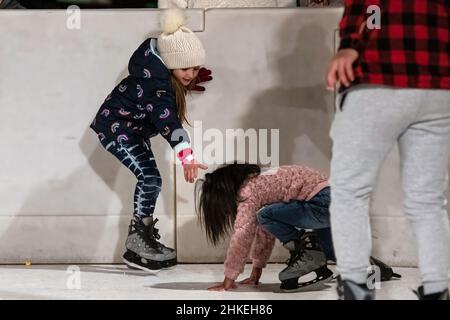 Prattville, Alabama, USA - 23. Dezember 2021: Ein junges Mädchen reicht ihre Hand aus, um einem anderen Mädchen zu helfen, das auf der Eislaufbahn in Histo gefallen ist Stockfoto