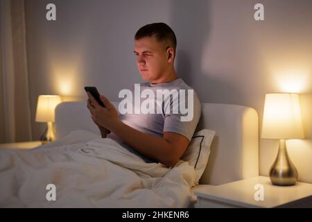 Junger Mann, der zu Hause mit dem Telefon im Bett sitzt Stockfoto