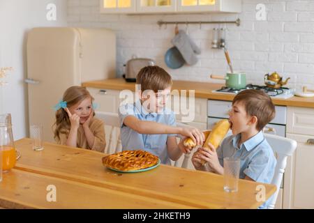 Fröhliche Kinder sitzen an einem Tisch in der Küche, spielen mit Laib Brot, während sie auf Essen warten. Zwei Brüder und Schwester haben Spaß beim Abendessen zu Hause. Familie verbringt am Wochenende Zeit zusammen zu Hause Stockfoto